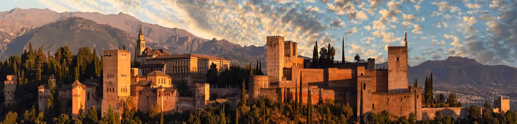 Türme und Mauern der Alhambra leuchten im warmen Abendlicht über Granada, dahinter Berge und Wolken