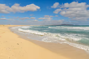 Ampia spiaggia sabbiosa deserta a Capo Verde, con onde turchesi e basse colline all'orizzonte sotto nuvole sparse
