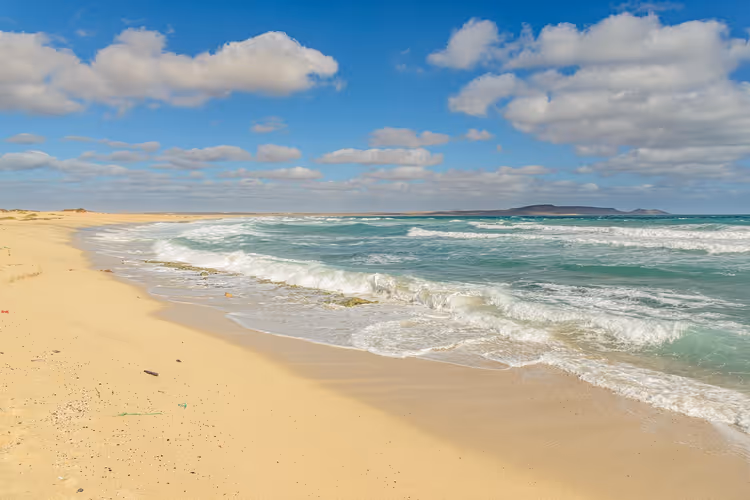 Breiter, leerer Sandstrand auf Kap Verde mit türkisfarbenen Wellen, flachen Hügeln am Horizont und vereinzelten Wolken