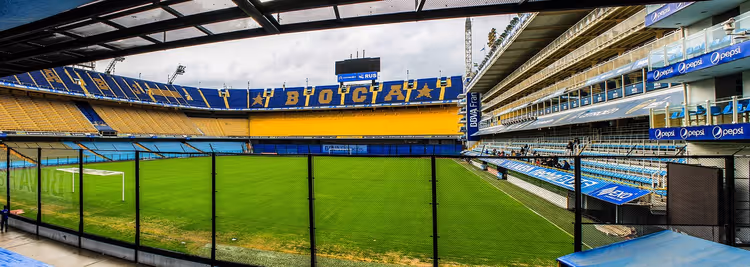 Leeres Fußballstadion mit blau-gelben Tribünen und grünem Rasen, durch einen Zaun gesehen, in Buenos Aires