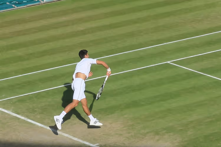 Tennisspieler in Weiß beim Aufschlag auf einem gestreiften Rasenplatz in Wimbledon, von der Tribüne aus im Sonnenlicht gesehen