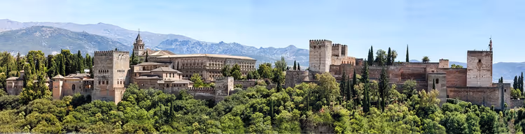 Festung und Paläste der Alhambra auf einem bewaldeten Hügel in Granada, dahinter Berge im Dunst