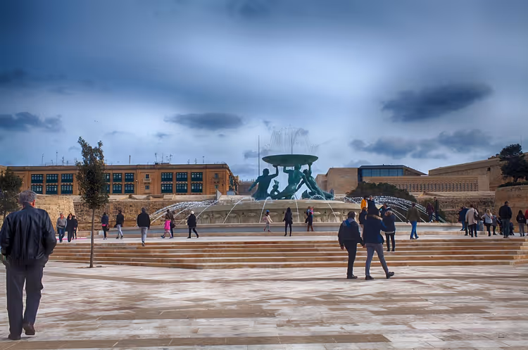 Menschen gehen über einen weiten gepflasterten Platz zum Tritonenbrunnen auf Malta unter dunklem Wolkenhimmel