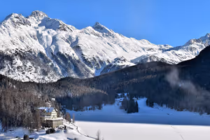 Zugefrorener, verschneiter See in St. Moritz mit Hotel am Ufer, dunklem Kiefernwald und sonnenbeschienenen Alpengipfeln