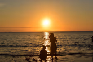 Sagome di due persone sulla battigia di una spiaggia che guardano un tramonto arancione su un mare calmo