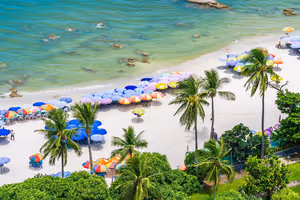 Vit sandstrand i Thailand kantad av färgglada parasoller och palmer, sedd uppifrån intill grunt grönt vatten