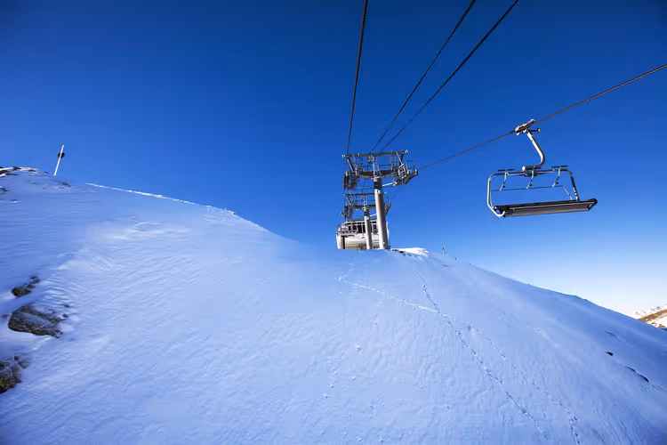 Leerer Sessel eines Sessellifts passiert eine Liftstütze über einem verschneiten Hang unter tiefblauem Himmel