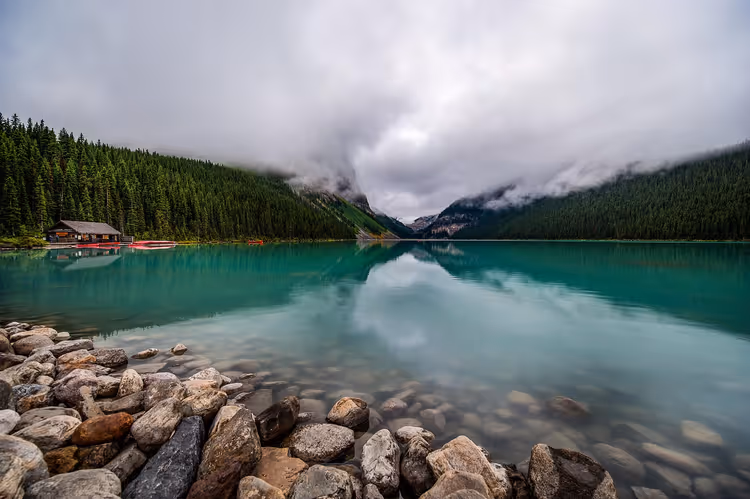 Türkisfarbenes Wasser des Lake Louise mit Felsblöcken am Ufer, einem Bootshaus und bewaldeten Hängen unter tiefen Wolken