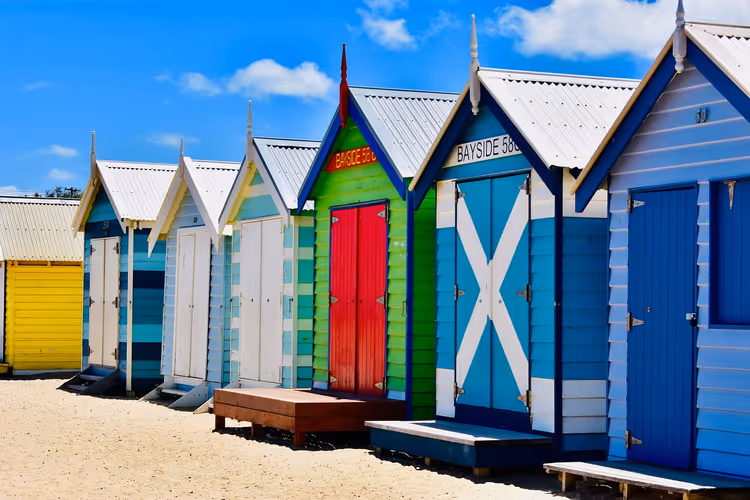 Reihe bunt gestrichener hölzerner Strandhäuschen in Blau, Grün, Rot und Gelb an einem Sandstrand bei sonnigem Himmel