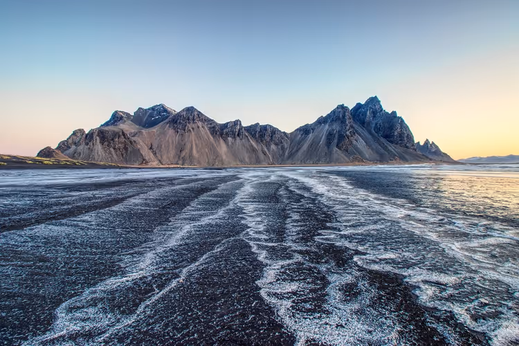 Zerklüfteter dunkler Bergkamm über einem schwarzen Sandstrand mit weißen Schaumstreifen in der Dämmerung