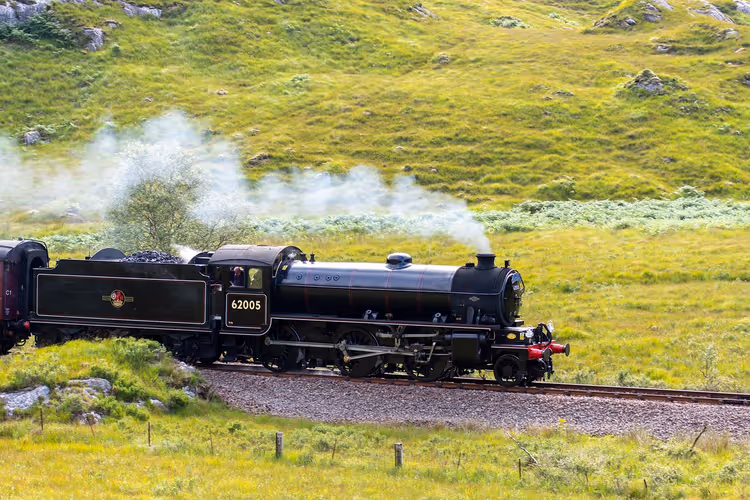 Locomotive à vapeur noire crachant une fumée blanche sur une voie traversant de vertes collines écossaises