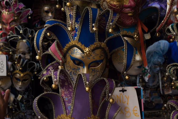 Karnevalsmasken im venezianischen Stil mit blauen und violetten Narrenkragen und Goldverzierung hängen an einem Marktstand