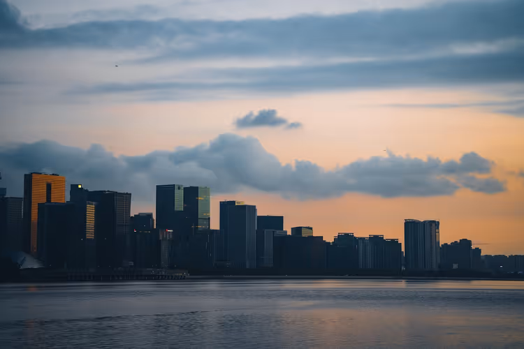 Silhouette einer Hochhaus-Skyline hinter ruhigem Wasser in der Abenddämmerung unter orangefarbenem Himmel mit grauen Wolken