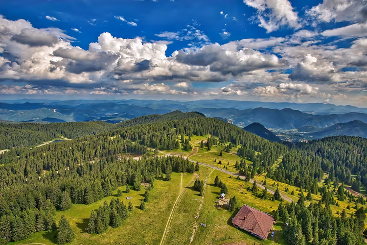 Luftaufnahme von fichtenbewachsenen Bergrücken, grünen Wiesen und einer Berghütte mit rotem Dach in Bulgarien unter Haufenwolken