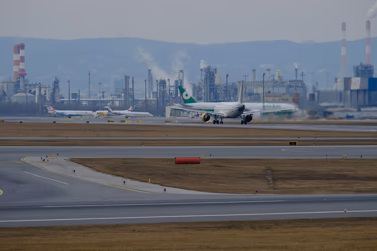 Passagierjet mit grünem Leitwerk auf einer Startbahn, dahinter abgestellte Flugzeuge und eine Raffinerie mit Schornsteinen im Dunst