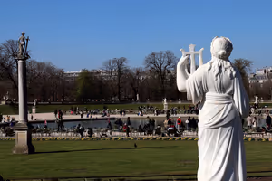 Weiße Marmorstatue über einem Teich und Menschen auf Stühlen in einem Pariser Garten unter klarem Himmel, die Bäume noch kahl