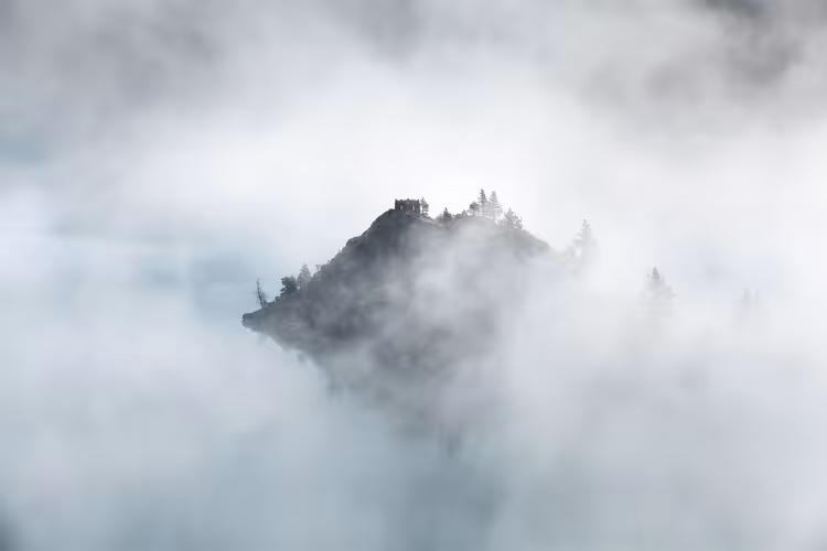 Felsige Bergkuppe mit Nadelbäumen und einem kleinen Gebäude ragt aus dichtem weißem Nebel