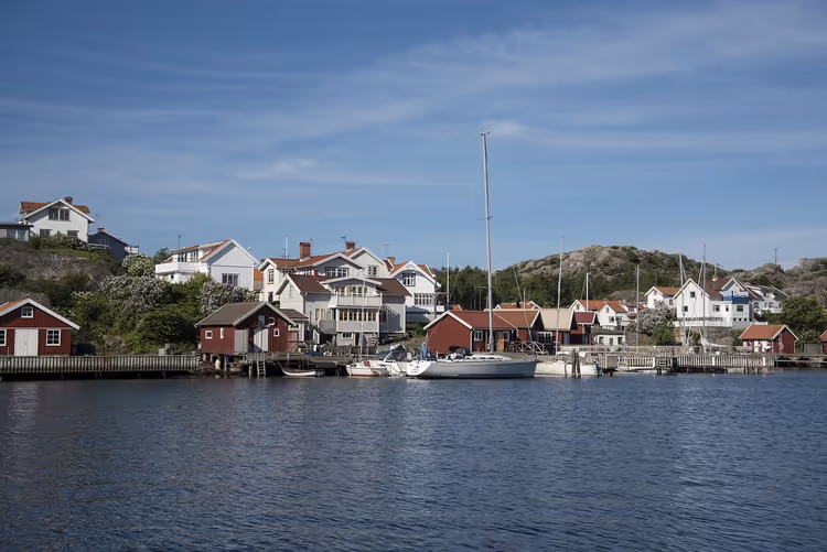 Rote Bootshäuser und weiße Holzhäuser an einem felsigen Ufer mit vertäuten Segelbooten in einem schwedischen Küstendorf