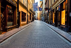 Leere gepflasterte Straße in der Stockholmer Altstadt mit ockerfarbenen Häusern, erleuchteten Schaufenstern und hängenden Bannern