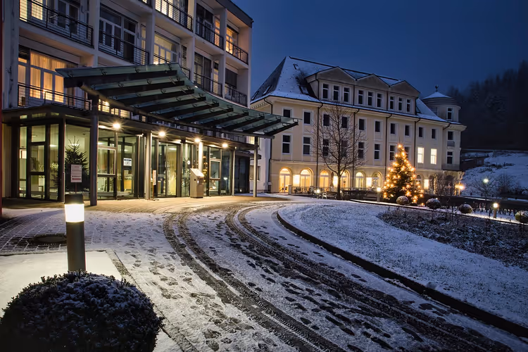 Hoteleingang mit Glasvordach und beleuchtetem Weihnachtsbaum an einem verschneiten Abend, Reifenspuren in der Zufahrt