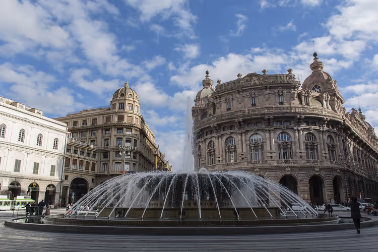 Großer runder Brunnen auf der Piazza De Ferrari in Genua, umgeben von reich verzierten Kuppelpalästen unter blauem Wolkenhimmel