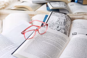 Lesebrille mit rosa Fassung auf einem Stapel aufgeschlagener Bücher, eines davon mit einer schwarz-weißen Stichillustration