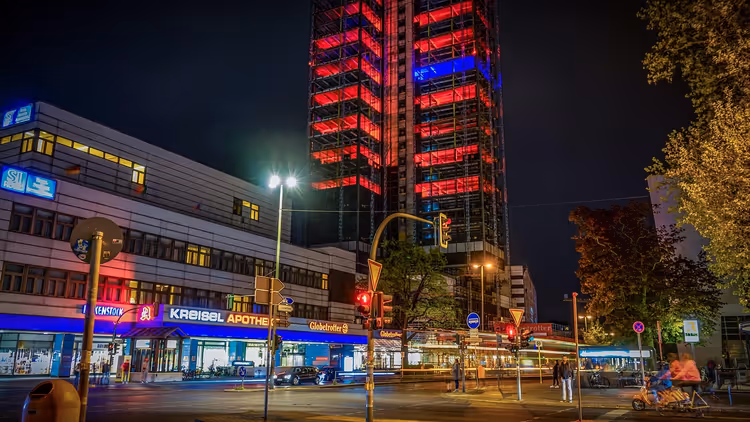 Rot und blau gestreift beleuchtetes Hochhaus über einer Straßenecke mit Läden und Ampeln bei Nacht in Berlin