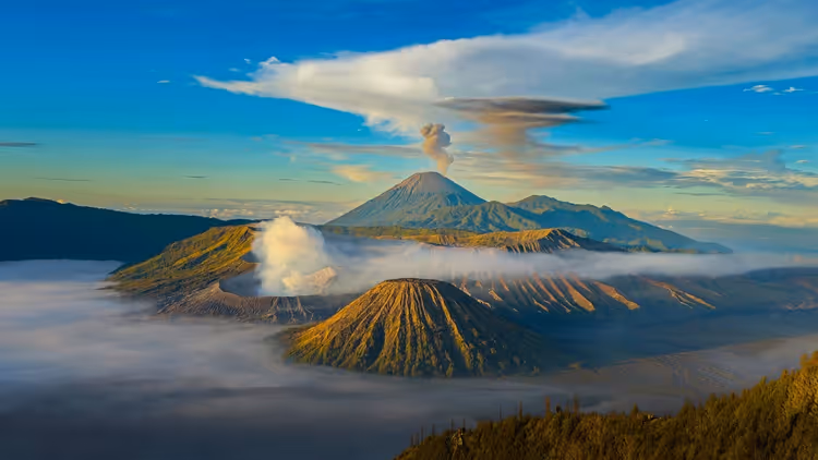 Vulkankegel ragen bei Sonnenaufgang in Indonesien aus einem Nebelmeer, aus einem Krater steigt eine Rauchfahne