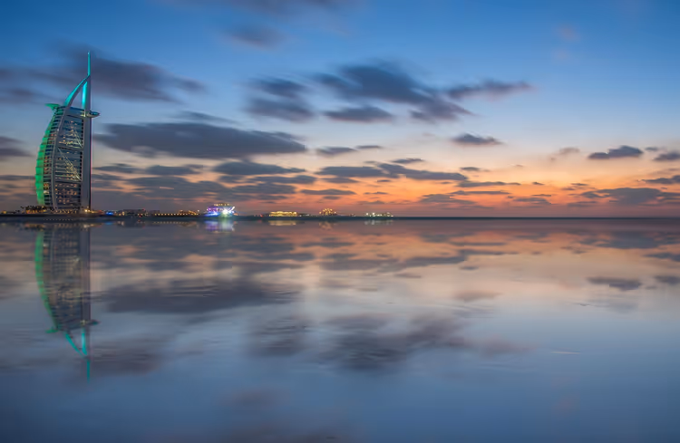 Segelförmiges Hotel Burj Al Arab in Dubai, in der Dämmerung grün beleuchtet, spiegelt sich im stillen Wasser unter orange-blauem Himmel