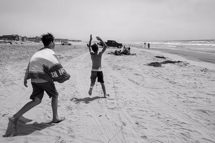 Schwarz-Weiß-Aufnahme: zwei Jungen rennen barfuß über einen weiten Sandstrand in Kalifornien, Pick-ups parken nahe der Brandung