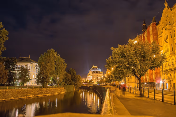 Nachts beleuchtetes Nationaltheater in Prag am Ende einer baumgesäumten Moldaupromenade, Lichter spiegeln sich im Wasser