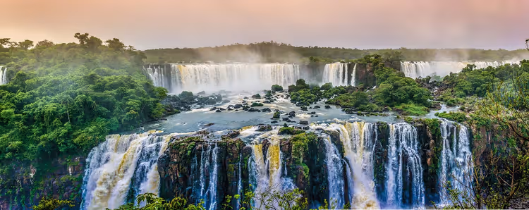 Iguazú-Wasserfälle stürzen in zwei Stufen über dunkle Felsen, umgeben von grünem Regenwald unter rosa dunstigem Himmel