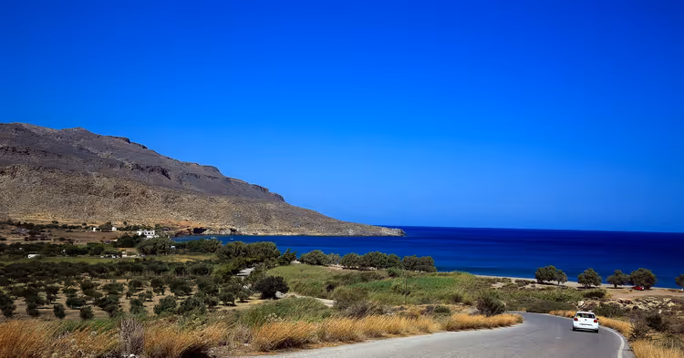 Weißes Auto auf einer Küstenstraße auf Kreta fährt an Olivenhainen vorbei, ein kahler Berg fällt zum tiefblauen Meer ab