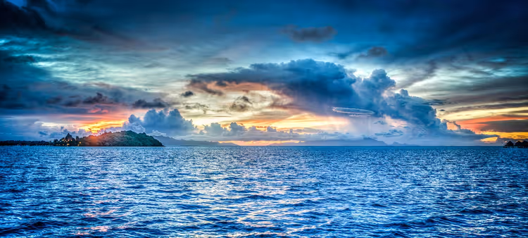 Dramatische wolken bij zonsondergang boven een rimpelende blauwe zee met een klein bebost eiland aan de horizon