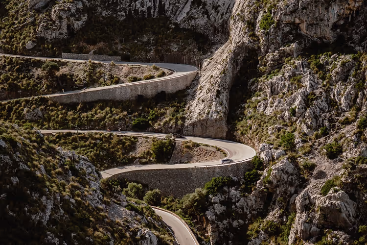 Schmale Straße schlängelt sich in Haarnadelkurven einen steilen, felsigen Berghang mit spärlichem grünem Gestrüpp auf Mallorca hinab