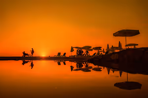Silhouetten von Sonnenschirmen, Liegen und Menschen, gespiegelt im stillen Wasser unter einem tieforangen Sonnenuntergang