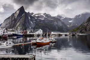 Fischerboote in einem ruhigen Hafen unter zerklüfteten Berggipfeln mit Schneestreifen und schweren Wolken