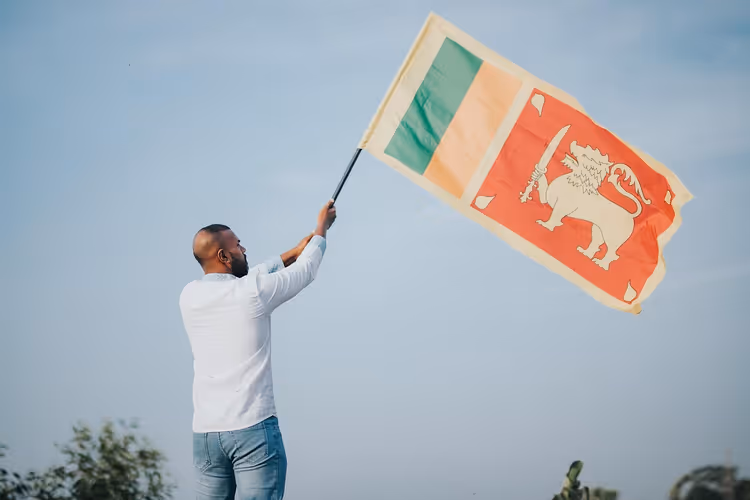 Mann in weißem Hemd und Jeans von hinten, der eine große Flagge Sri Lankas vor blassblauem Himmel schwenkt