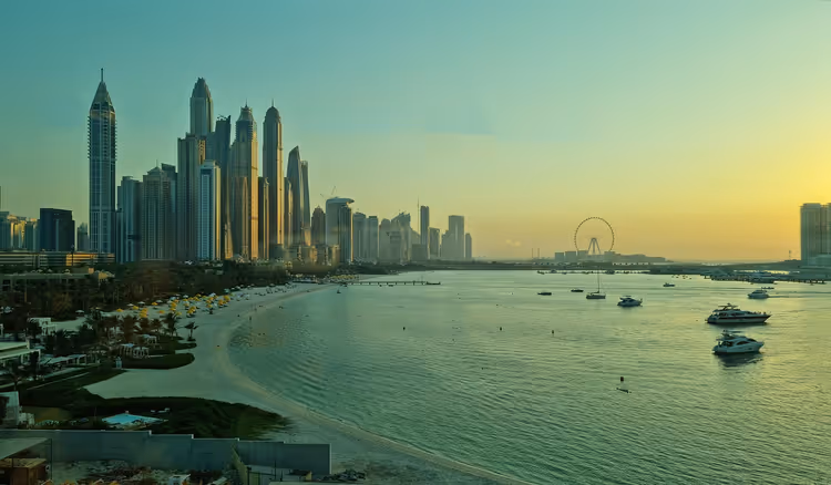 Uferfront von Dubai bei Sonnenuntergang mit Wolkenkratzern, geschwungenem Sandstrand, Yachten auf ruhigem Wasser und Riesenrad