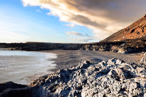 Bucht mit schwarzem Sand auf La Palma, gesäumt von rauem Lavagestein und einem rötlichen Vulkanhang unter tiefen, sonnenbeschienenen Wolken