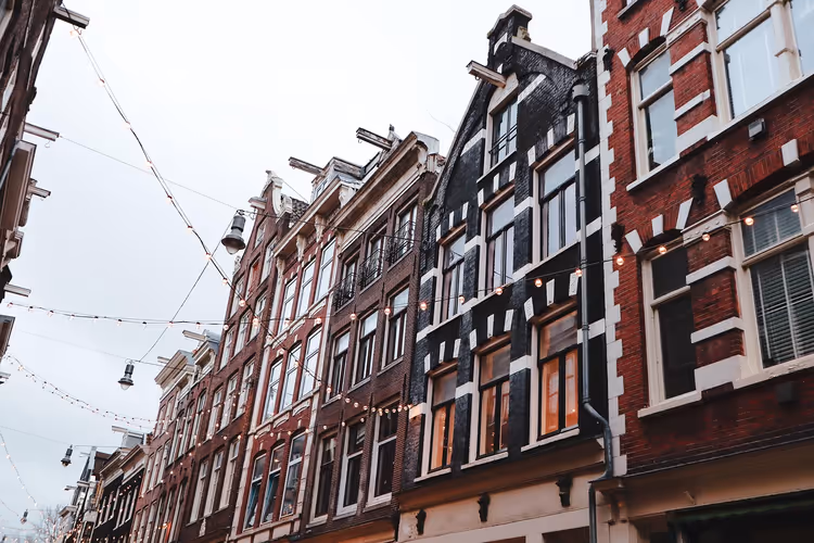 Reihe schmaler Stadthäuser aus dunklem und rotem Backstein mit hohen Fenstern und Lichterketten über einer Straße in Amsterdam