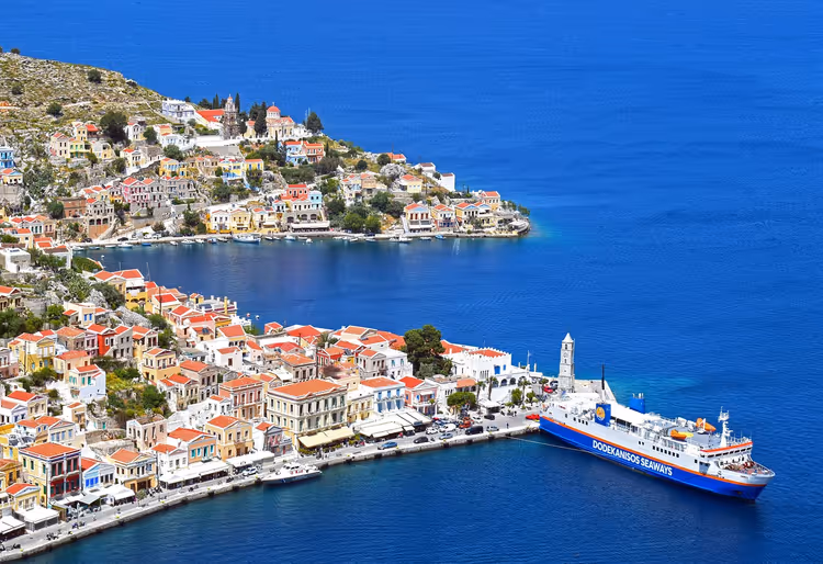 Blau-weiße Fähre neben einem Uhrturm im Hafen einer griechischen Insel, gesäumt von pastellfarbenen Häusern mit roten Dächern