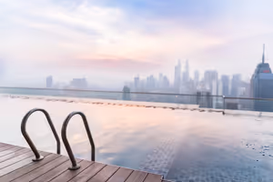 Infinity-Pool auf dem Dach mit Metallleiter und Holzdeck, Blick auf eine dunstige Wolkenkratzer-Skyline in der Dämmerung