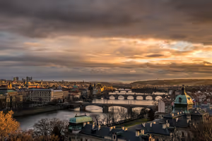 Reihe von Brücken über die Moldau in Prag unter schweren Wolken, die die tiefe Sonne orange färbt, vorn grüne Kupferkuppeln