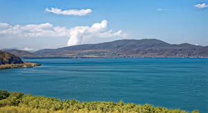 Lago di montagna turchese in Armenia con un villaggio sulla sponda opposta, colline brune e spoglie e arbusti verdi in primo piano