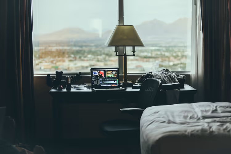 Halbdunkles Hotelzimmer mit Laptop und Lampe auf einem Schreibtisch an einem breiten Fenster mit Blick auf Stadt und Berge im Dunst