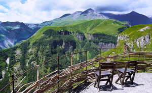Zwei Holzbänke an einem Aussichtspunkt mit Flechtzaun, Blick auf grüne Hänge und Felsgipfel des Kaukasus in Georgien