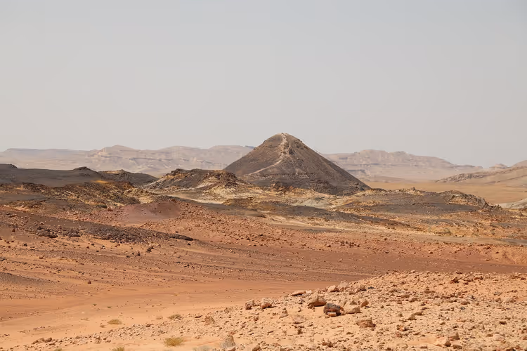 Dunkler kegelförmiger Hügel über einer rötlichen felsigen Wüstenebene in Israel, in der Ferne Bergrücken im Dunst