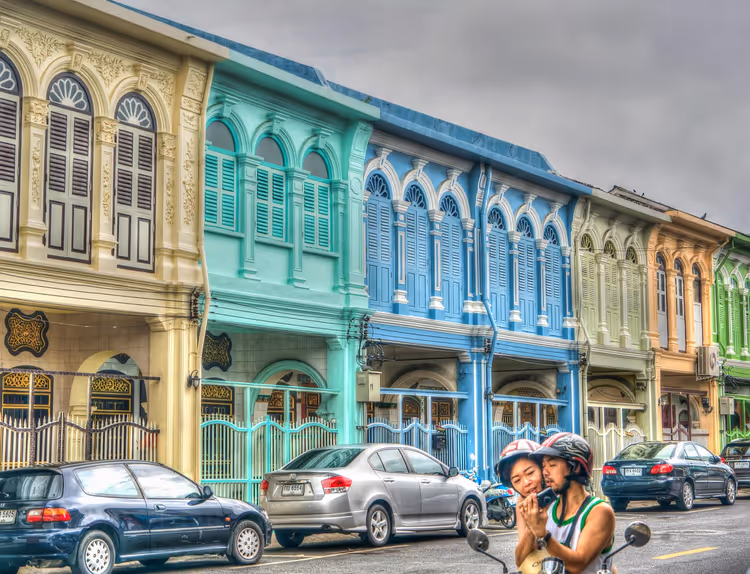 Pastellgelbe, türkise und blaue Ladenhäuser mit Bogenfensterläden an einer Straße in Phuket Town, davor Rollerfahrer
