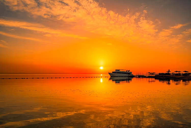 Jacht an einem Steg mit Sonnenschirmen bei Sonnenaufgang, oranger Himmel spiegelt sich im ruhigen flachen Meer in Hurghada